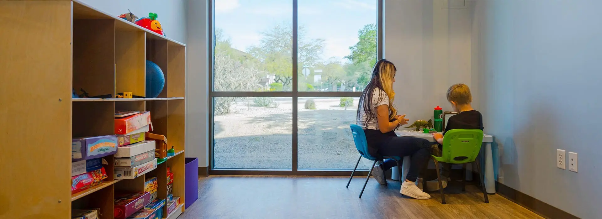 A woman and child play with toys in the corner of a room while the opposite wall holds a bookshelf of toys and games