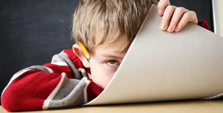 a little boy at school with a pencil behind his ear hiding behind a notebook