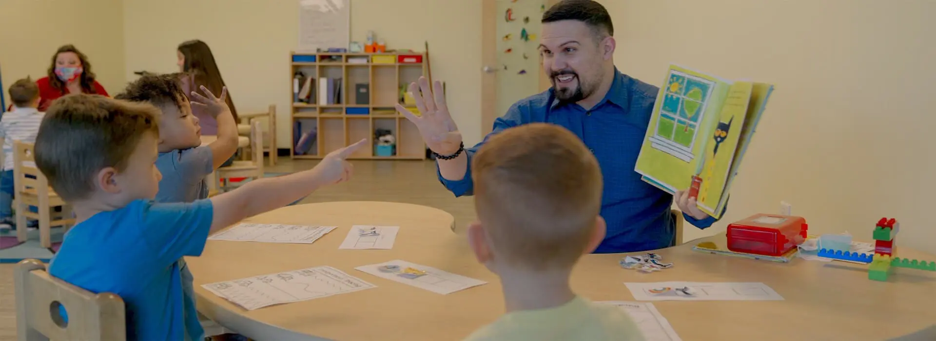 a man holds up the number 5 while holding a book while one boy is pointing at him and another is sitting